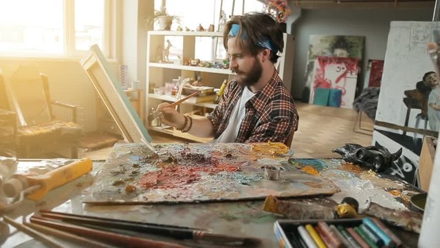 Bearded talented painter creating a picture in huge spacious studio, wearing casual checked t-shirt as working at the desk full of art equipment, indoor shot on sunny morning