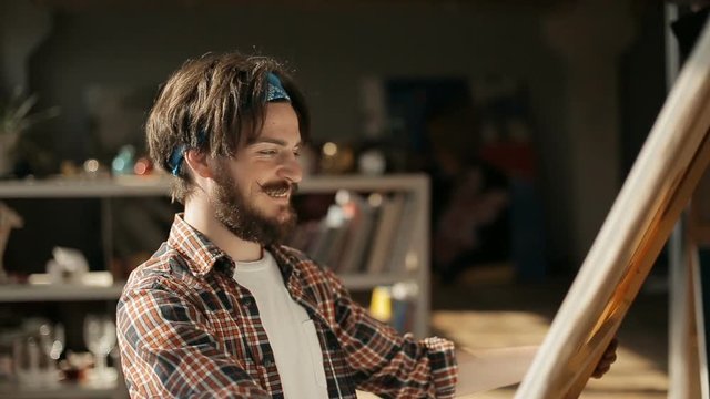 Smiling inspired artist looking at canvas, examing it carefully before hanging the picture on wall, wearing checked t-shirt, white shirt and blue bandana while working in studio