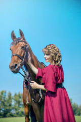 Obraz premium Beautiful girl in a long red dress stands next to the brown horse. A girl stroking a horse.