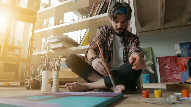 Concentrated caucasian painter creating picture as sitting on the floor, preparing for art exhibition in light studio filled with modern paintings