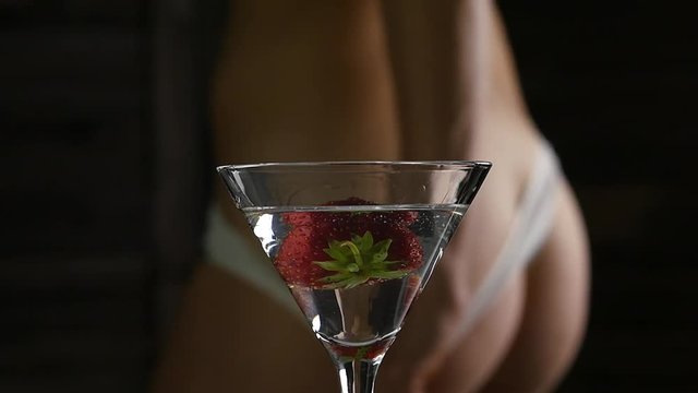 cocktail with strawberry and blured female buttocks in white panties in a dark background. dating and party concept. slow motion