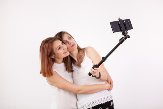 Portrait of two beautiful young woman taking a selfie with a selfie stick and making different funny and silly faces while doing it. Studio photo over a white wall