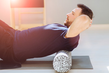 Handsome man performing back exercise on a foam roller being assisted at pilates studio. Patient...