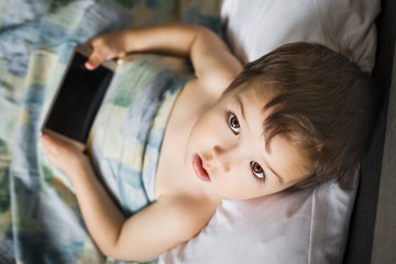 young brainy modern boy holding touching mobile phone lying on bed in morning