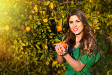 Young woman, outdoors at sunset in a orange orchard, looking at camera and smiling, holding an orange fruit. Healthy lifestyle concept, skin and hair care concept.