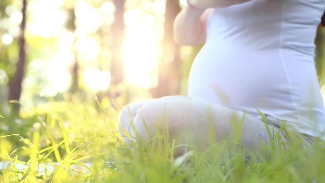 Close-up on belly of young healthy pregnant woman doing yoga in natureon summer park, outdoors at sunset