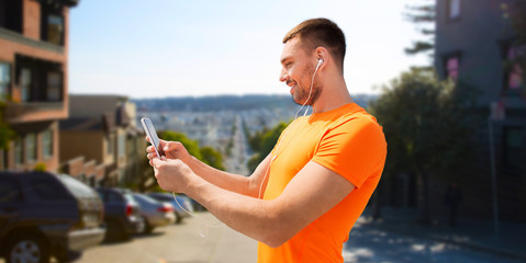 fitness, sport and technology concept - smiling young man with smartphone and earphones listening to music over san francisco city background