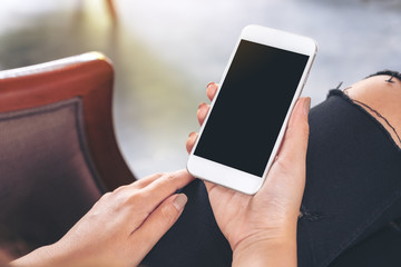 Mockup image of woman's hands holding white mobile phone with blank black desktop screen while sitting in cafe