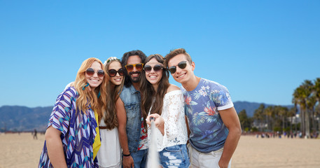 tourism, travel and summer holidays concept - smiling young hippie friends taking picture by selfie stick over venice beach background in california