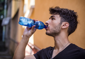 Handsome young man drinking water from plastic bottle, standing in European city street, leaning against a wall