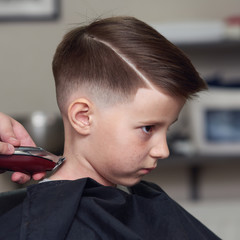 Barber is shaving a hair to Caucasian boy in barbershop. - 212587568