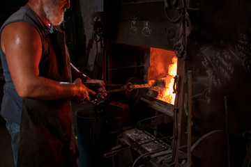 The smith artisan in his apron heats the workpiece in the burning furnace in the smith