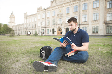 Portrait of a student sitting on a grass at a university campus and reading a book. Man with a beard studies outdoor in the background of the university building.