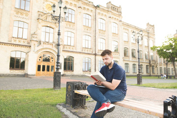 Fototapeta na wymiar Concentrated student sitting on the background of the university and reading a book. Man with a beard sits on a bench in the campus of the university, reads a book and learns. Learning the concept