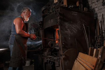 The smith artisan in his apron heats the workpiece in the burning furnace in the smith