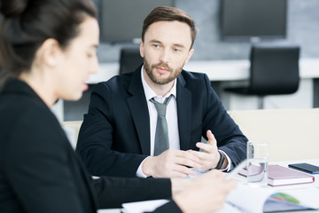 Portrait of successful handsome businessman talking to partner while sitting at meeting table in conference room, copy space