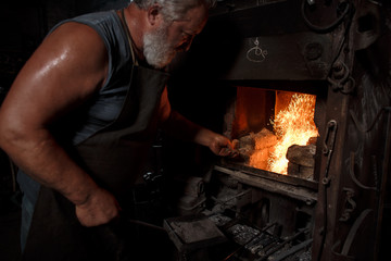 The smith artisan in his apron heats the workpiece in the burning furnace in the smith