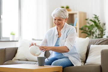age, drink and people concept - senior woman pouring tea from teapot to cup and drinking it at home