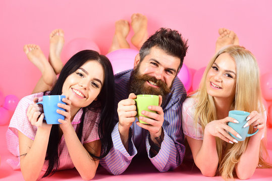 Lovers drinking coffee in bed. Lovers in bed concept. Man and women, friends on smiling faces lay, pink background. Man and women in domestic clothes, pajamas. Threesome relax in morning with coffee.