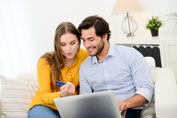 cheerful young couple on sofa at home websurfing and shopping online with credit card, a laptop computer and internet technology