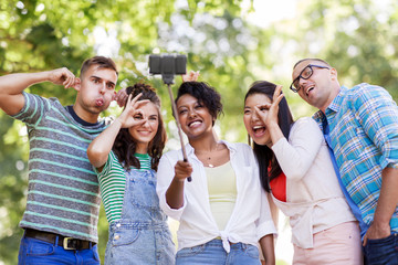 people, friendship and international concept - happy smiling young woman and group of happy friends taking picture by selfie stick in park