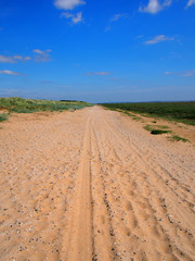 a straight long dry sand road with tire tracks and footprints extending to the horizon surrounded by grass covered dunes with a bright blue summer sky