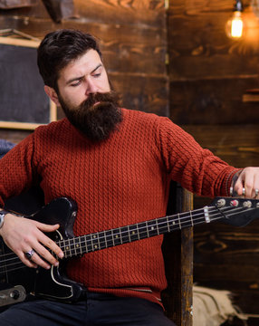 Rock musician with brutal look posing with instrument. Bearded man tuning electric guitar. Man with stylish beard and mustache in terracotta knitted sweater and black jeans relaxing, music concept