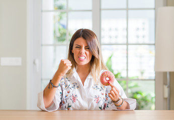 Young woman at home eating a doughnut annoyed and frustrated shouting with anger, crazy and yelling with raised hand, anger concept