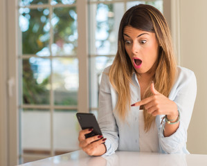 Young woman holding smartphone surprised. Panic and shock emotion at home