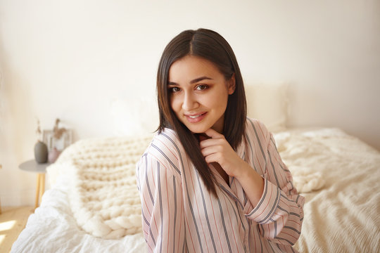 Portrait of charming cute young dark haired female in silk night suit with stripes spending lazy slow morning in bedroom, having happy carefree facial expression. People, rest and relaxation