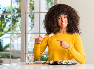 African american woman eating sushi using chopsticks at home very happy pointing with hand and...