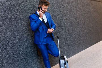 Portrait of handsome young man on business trip with his luggage talking on phone at airport.