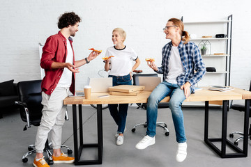 happy young coworkers eating pizza and smiling each other in office