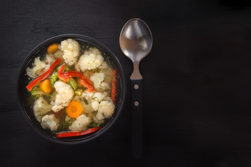 Vegetable soup of cauliflower, carrots, tomato, pepper and herbs in a black plate on a black background with a spoon. Copy space. Top view