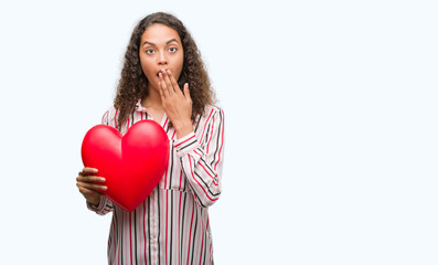 Young hispanic woman in love holding red heart cover mouth with hand shocked with shame for mistake, expression of fear, scared in silence, secret concept