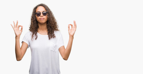 Young hispanic woman wearing sunglasses relax and smiling with eyes closed doing meditation gesture with fingers. Yoga concept.