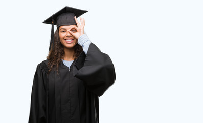 Young hispanic woman wearing graduation uniform with happy face smiling doing ok sign with hand on eye looking through fingers