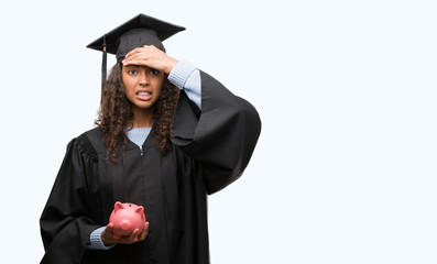 Young hispanic woman wearing graduation uniform holding piggy bank stressed with hand on head,...