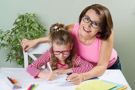 Mom Helps The Child Write. Girl 6, 7 Years Old With Glasses Sitting At The Table And Writing In Notebook