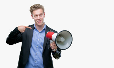 Young handsome blond man holding a megaphone with surprise face pointing finger to himself