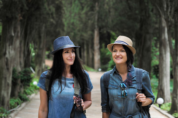 Two young beautiful brunette girls in jeans dresses and hats walk along the road of park with tall green trees.