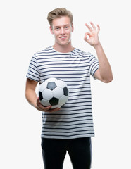 Young handsome blond man holding soccer ball doing ok sign with fingers, excellent symbol