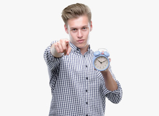 Young handsome blond man holding alarm clock pointing with finger to the camera and to you, hand...