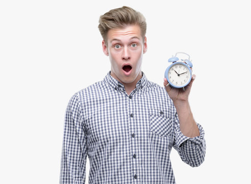 Young handsome blond man holding alarm clock scared in shock with a surprise face, afraid and excited with fear expression