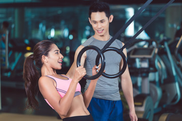 Side view of a fit and cheerful young woman doing ring biceps curl exercise during functional training workout with a professional personal trainer 