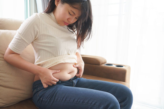 Woman holding her own belly when sitting down on sofa in the living room.