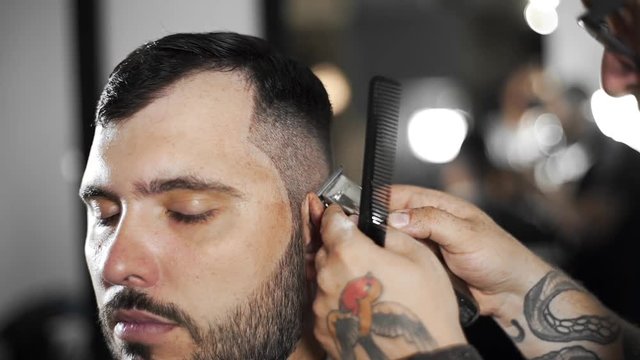 Tattoed barber makes haircut for customer at the barber shop by using hairclipper, man's haircut and shaving at the hairdresser, barber shop