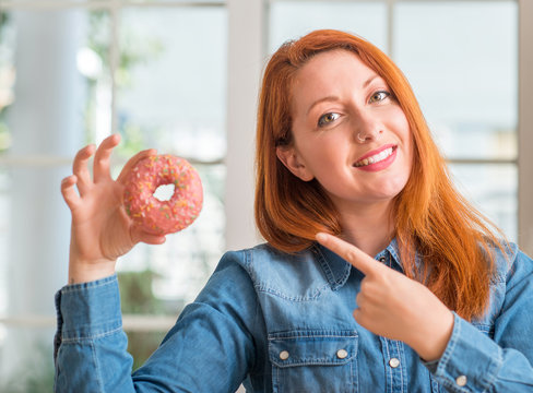 Redhead woman holding donut at home very happy pointing with hand and finger to the side