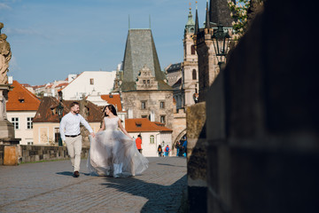 Happy wedding couple of groom and bride running holding hands on the Charles bridge on sunny day....