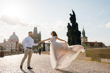 Beautiful happy wedding couple of groom and bride in gorgeous dress dancing and smiling while walking on the Charles bridge i the evening on sunset. Prague, Czech Republic